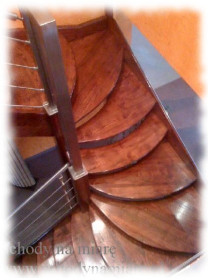 Spiralne schody z drewna o zaokrąglonych stopniach, z metalową balustradą z cienkimi, poziomymi prętami, widok z góry.